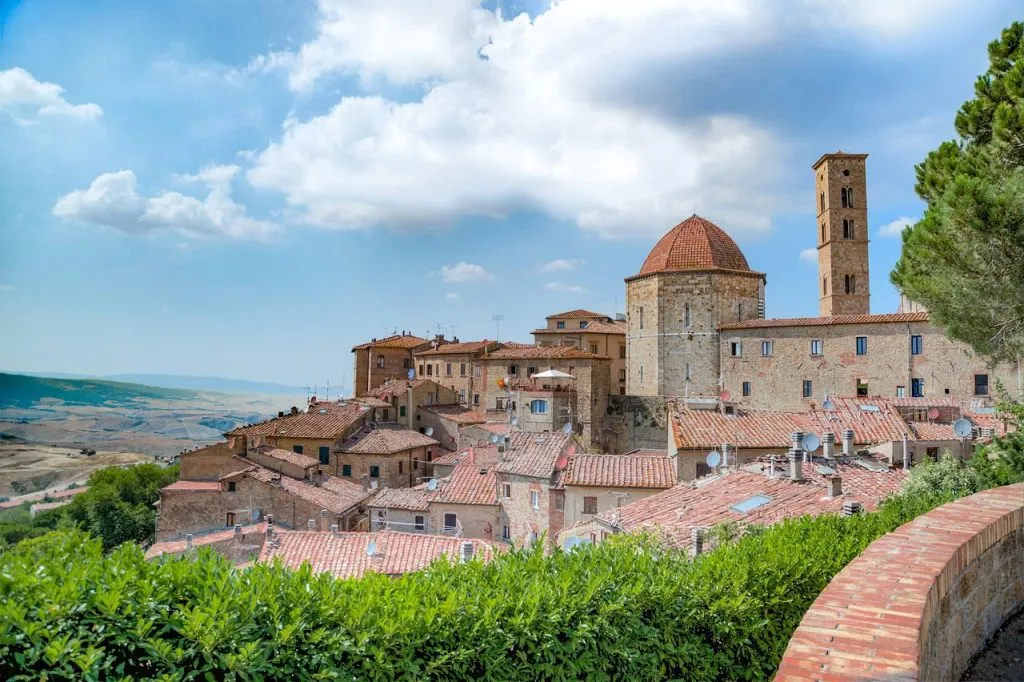 Town in Tuscany with classic European architecture, a low foreground hedge, and vast, dry plains visible on the horizon