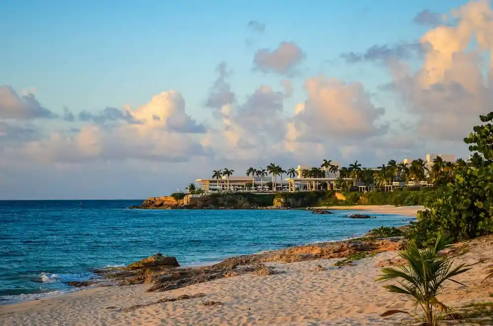 The shoreline of Anguilla with gentle waves, light-colored sand, and white buildings atop a low cliff
