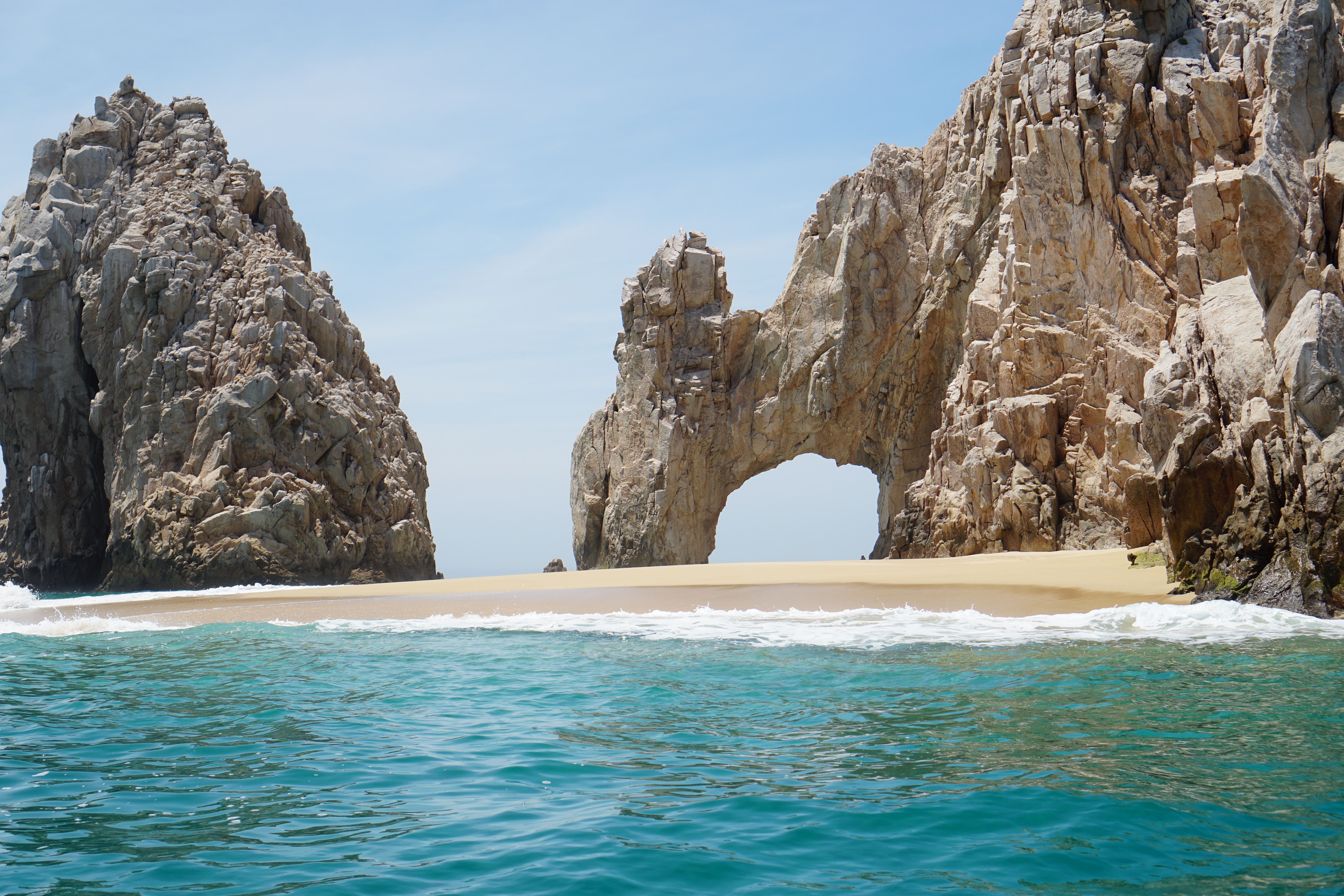 A high, rugged rock arch next to a sheer cliff face, with a patch of golden sand at the base