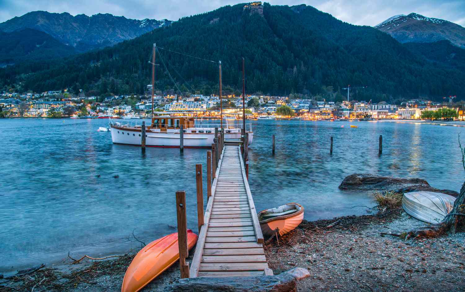 Evening view of Queenstown, New Zealand, showing a moored boat, small rowboats on the shore, and glowing town lights
