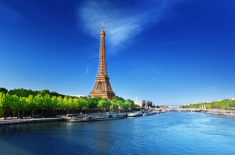 A clear, sunny day in Paris with the Eiffel Tower dominating the skyline above a tree-lined riverbank