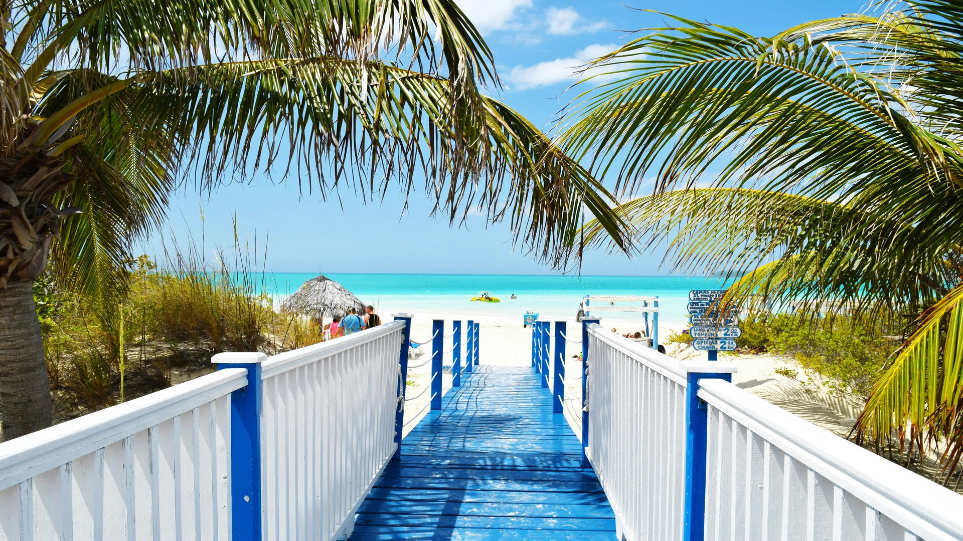 A bright blue and white wooden walkway leads down to a sunny white-sand beach and the turquoise ocean
