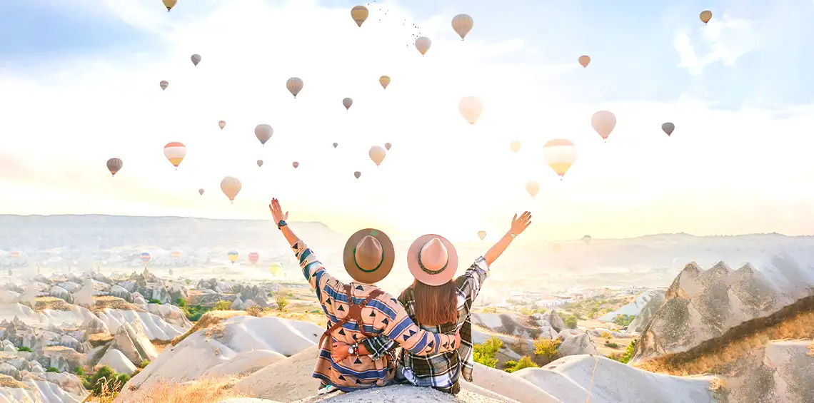 A couple sitting on a high hill with their arms raised, watching dozens of colorful hot air balloons floating over the unique landscape of Cappadocia, Turkey