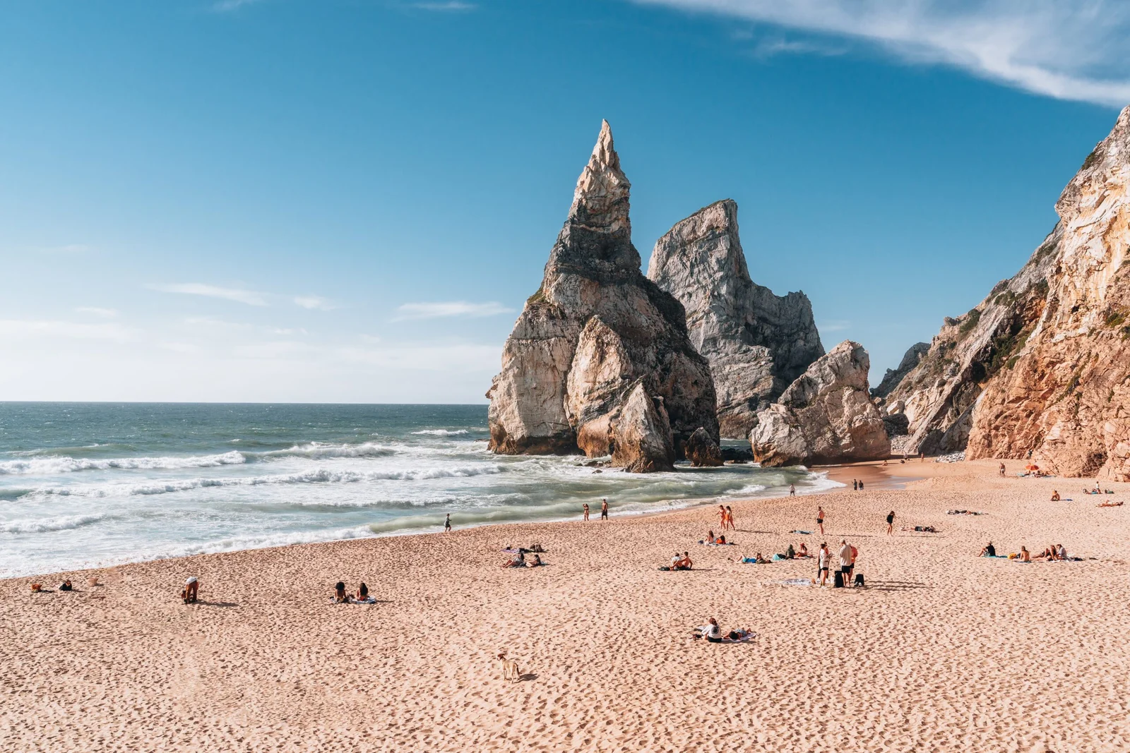A wide, sandy beach in Portugal with people sunbathing, backed by massive, dramatic sea stacks and cliffs under a clear blue sky
