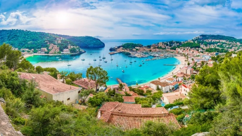 High view of a Spanish coastal village, a large harbor filled with boats, and red-roofed buildings under a blue, cloudy sky