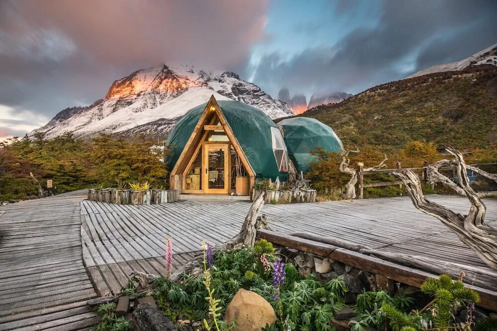 A remote eco-camp in Patagonia with a wooden path leading to a dome structure, surrounded by dramatic mountains and cloudy sky