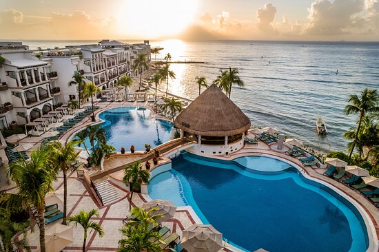 Sunrise over a coastal resort showing the blue swimming pool, lounge chairs, and an arching white hotel building