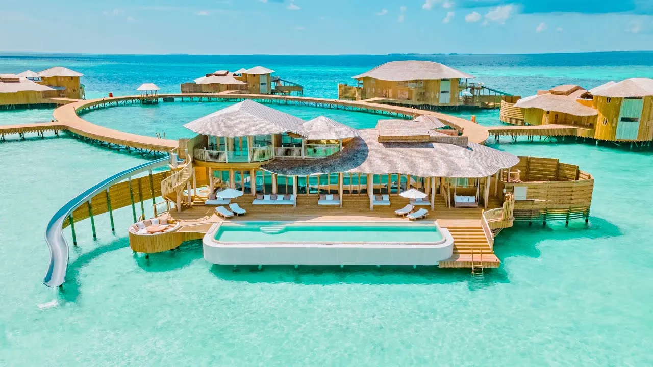 Maldives resort complex with wooden decks, pools, and slides, contrasting with the pale blue and green ocean water