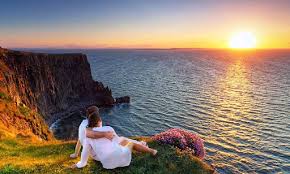 A wide shot of a couple enjoying the view of a massive sea cliff and the ocean at sunset, with warm golden light