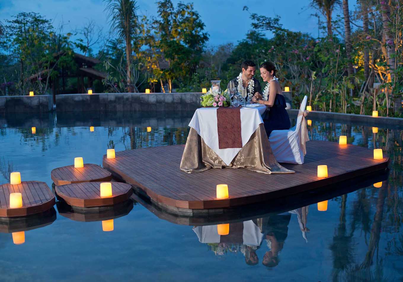 A couple enjoying a private candlelit dinner on a floating wooden deck in a pool at night
