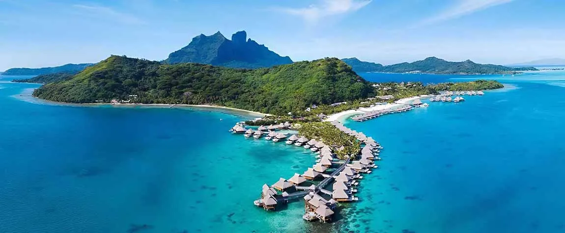 A remote island paradise with clear water, green trees, and many small wooden huts on stilts stretching out into the sea