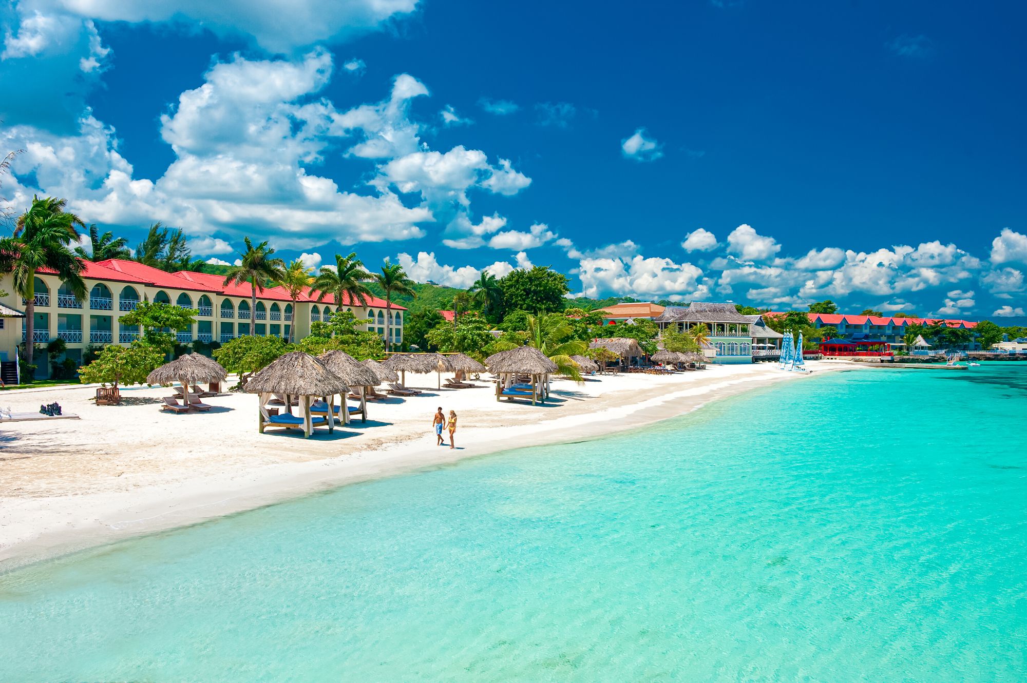 A couple walks on a white-sand beach next to bright turquoise water and a large resort in Jamaica