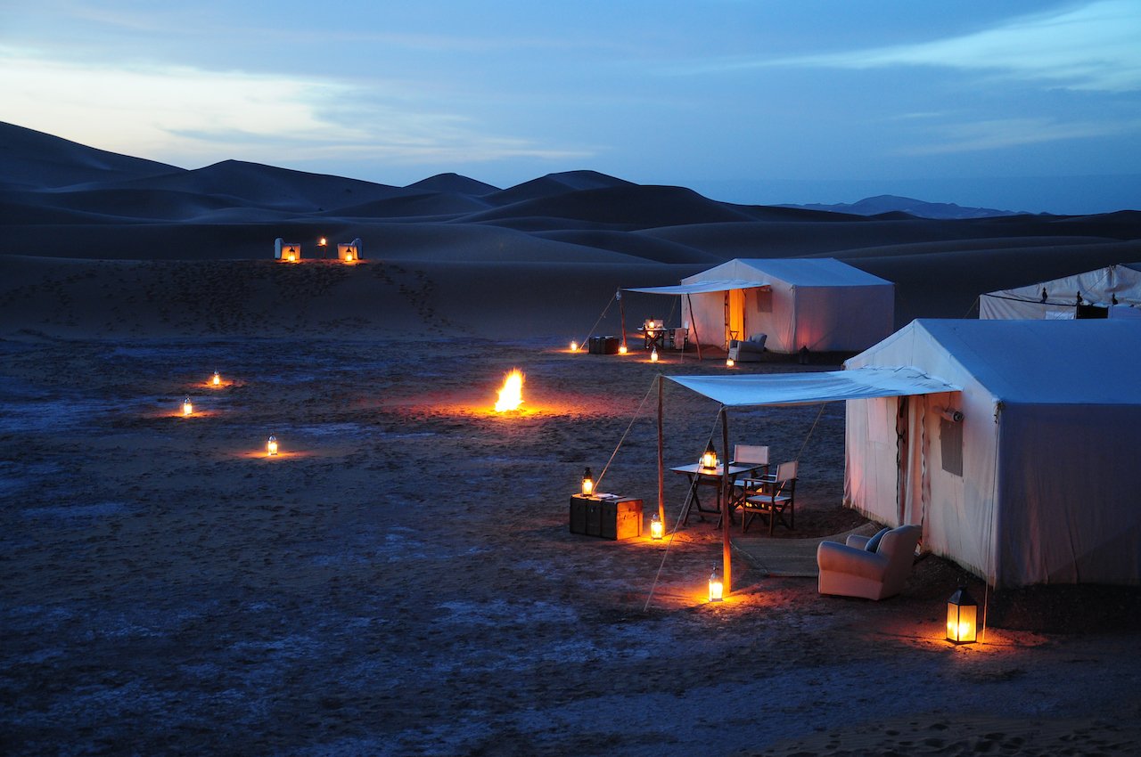 Luxury desert camp in Morocco at twilight, with white tents, lanterns, and a small campfire under dark sand dunes